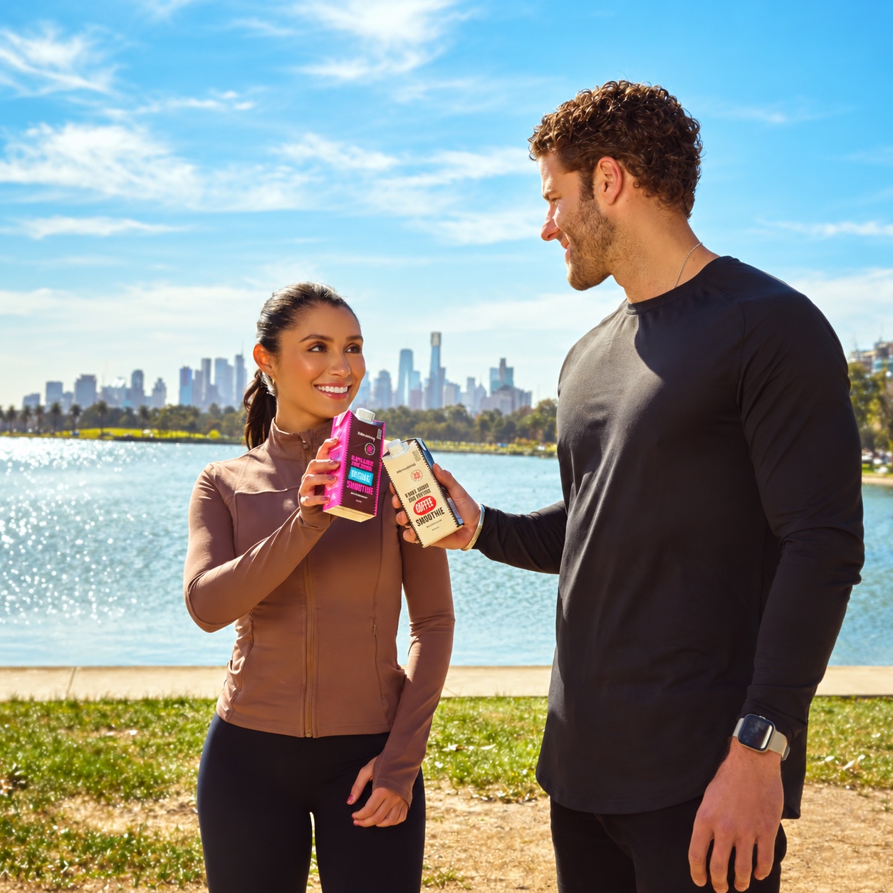 Two gym goers by the water with the Melbourne skyline behind them, holding 99th Monkey chocolate and coffee protein smoothies