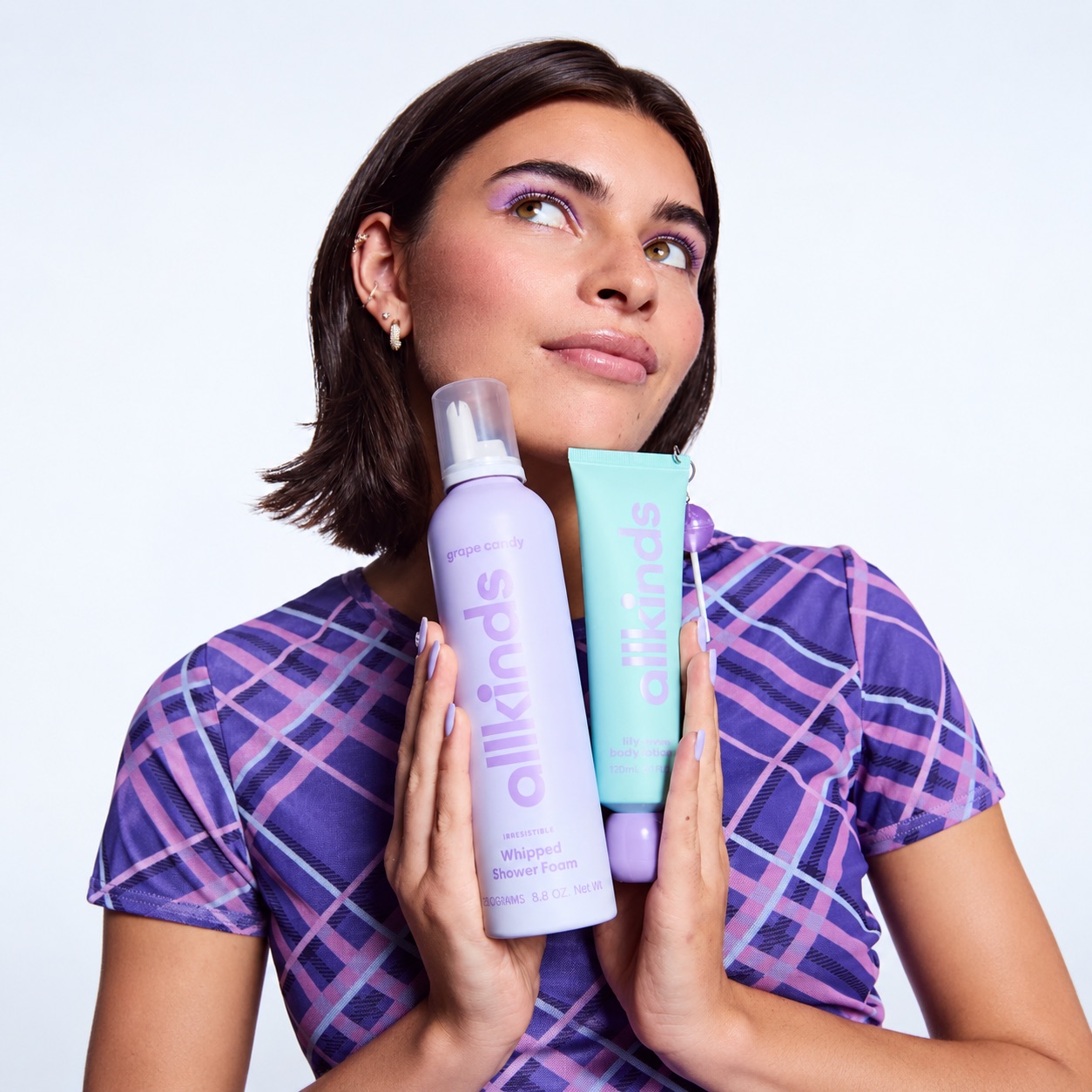 A model in a purple check tee holding Allkinds Grape Candy whipped shower foam and a mint body lotion to her cheek