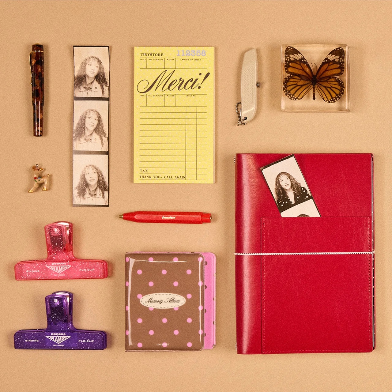 A Milligram flat lay on a tan background: a tortoiseshell fountain pen, a photo booth strip, a yellow Merci receipt pad, a framed monarch butterfly, a red Kaweco pen, glittery pink and purple binder clips, a polka dot memory album and a red leather notebook