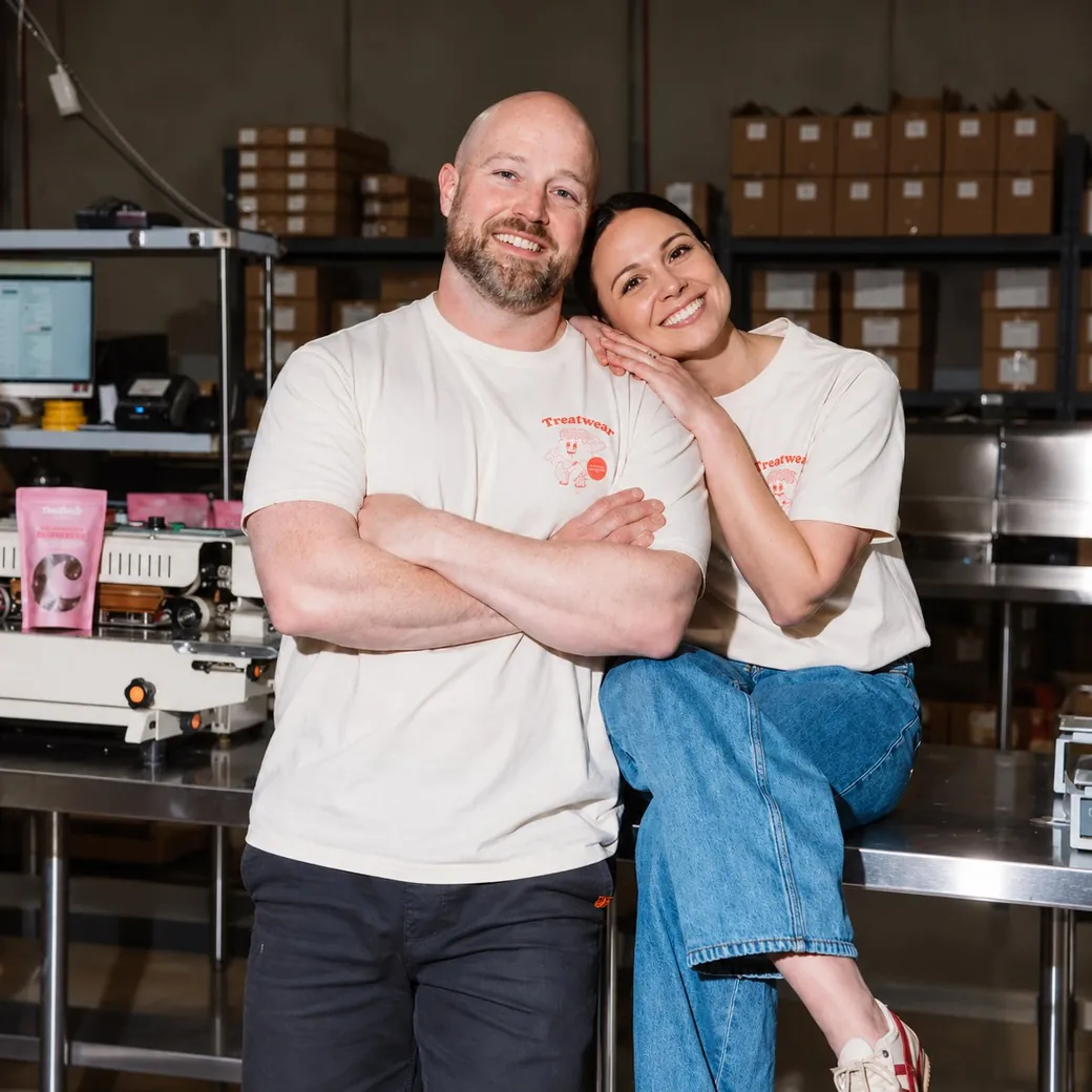 Matt and Tara Grisold in matching cream Treatwear tees, smiling in front of the packing bench and shelves of boxed stock in the Coated warehouse