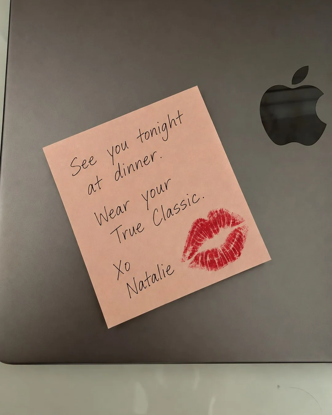 A pink sticky note with a lipstick kiss on a closed laptop: “See you tonight at dinner. Wear your True Classic. Xo Natalie”